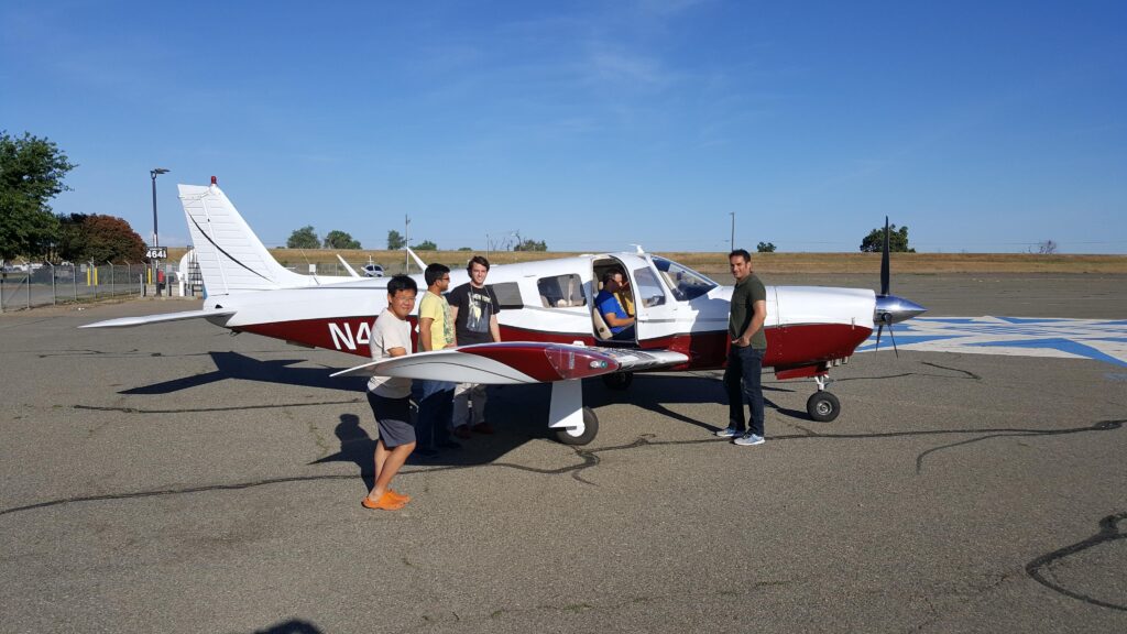 Four people stand around a small red and white airplane parked on an airfield under a clear blue sky. One person stands near the wing, while others are positioned near the door and tail section of the plane. - CFI Academy
