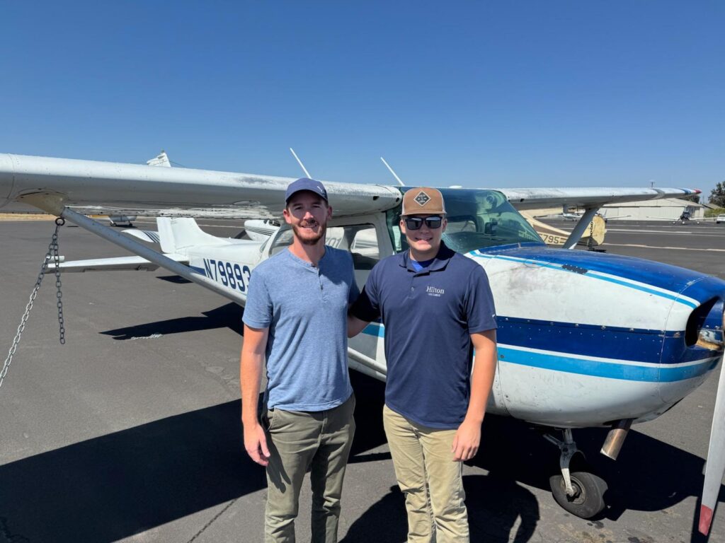 Two men stand side by side, smiling in front of a small white and blue single-engine airplane on a sunny day at an airport with a clear blue sky above. - CFI Academy