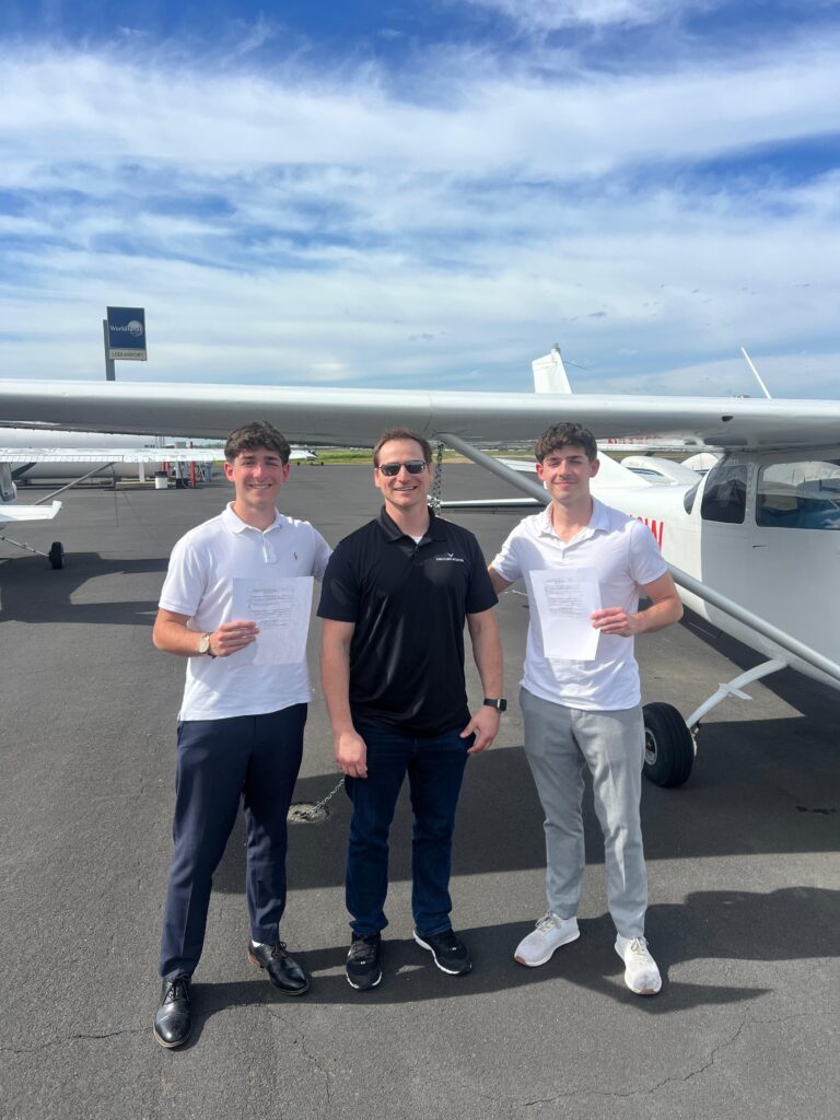 Three MEIs stand smiling in front of a small airplane on an airport tarmac, perhaps discussing the CFII MEI order. The two on the left and right hold papers, while the man in sunglasses and a black shirt stands center under a blue sky with clouds. - CFI Academy