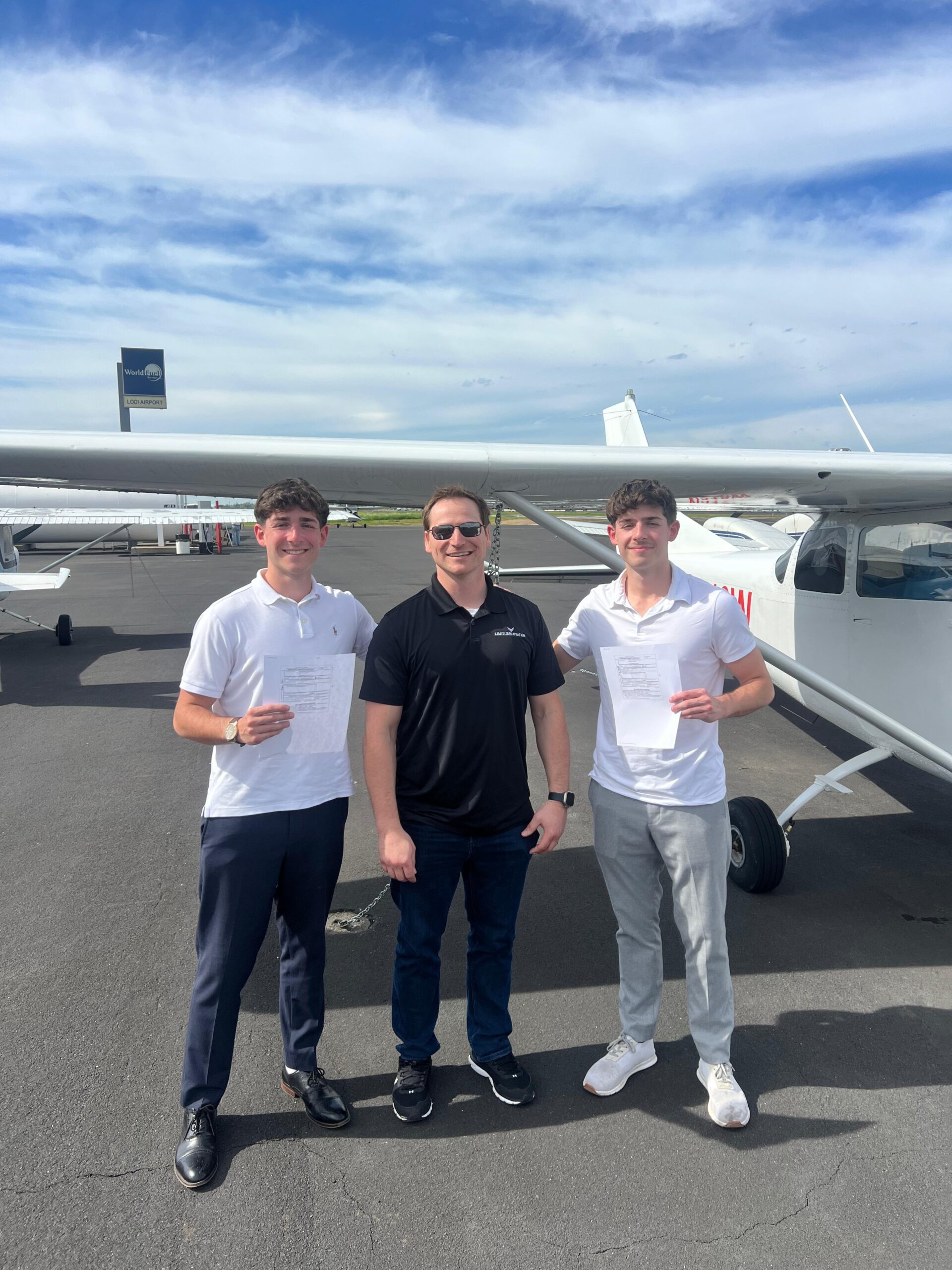 Three MEIs stand smiling in front of a small airplane on an airport tarmac, perhaps discussing the CFII MEI order. The two on the left and right hold papers, while the man in sunglasses and a black shirt stands center under a blue sky with clouds. - CFI Academy