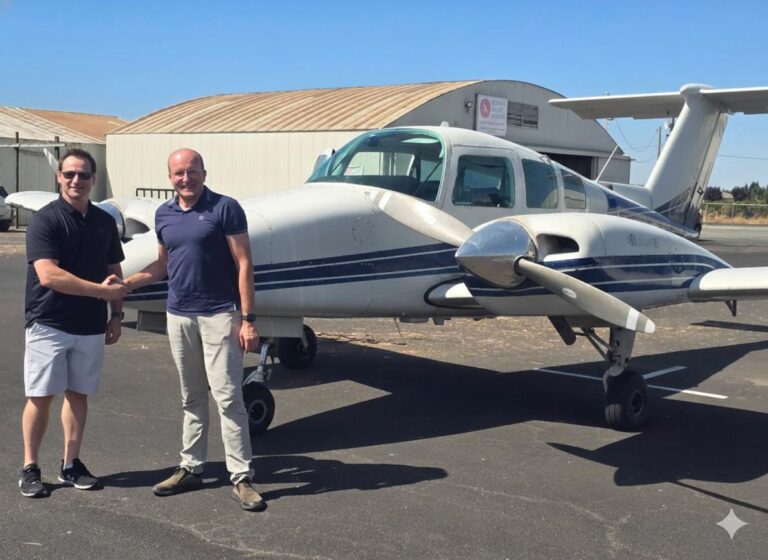 Two MEIs stand on an airport tarmac smiling and shaking hands in front of a small twin-engine airplane, celebrating the completion of an accelerated MEI course, with hangars and a clear sky in the background. - CFI Academy