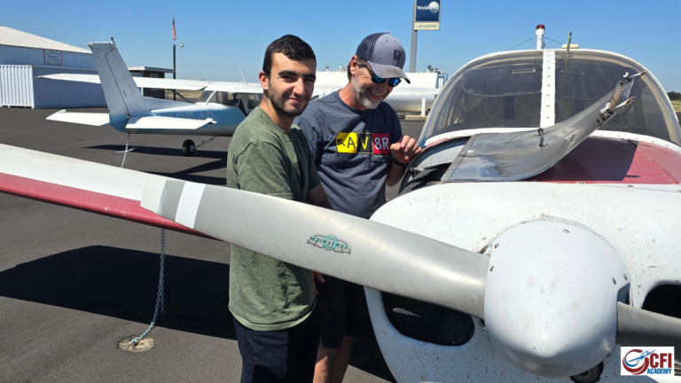 Two men stand beside a small propeller plane on an airport tarmac, possibly discussing accelerated CFI training. One wears a green shirt, the other a gray shirt and cap. A hangar and another plane are visible in the background. - CFI Academy