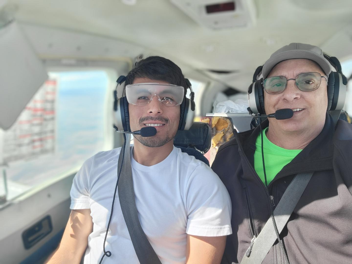 Two men wearing headsets and seatbelts sit in the cockpit of a small airplane. The man on the left wears a white shirt and fogged safety goggles, while the man on the right wears glasses, a cap, and a green shirt. - CFI Academy