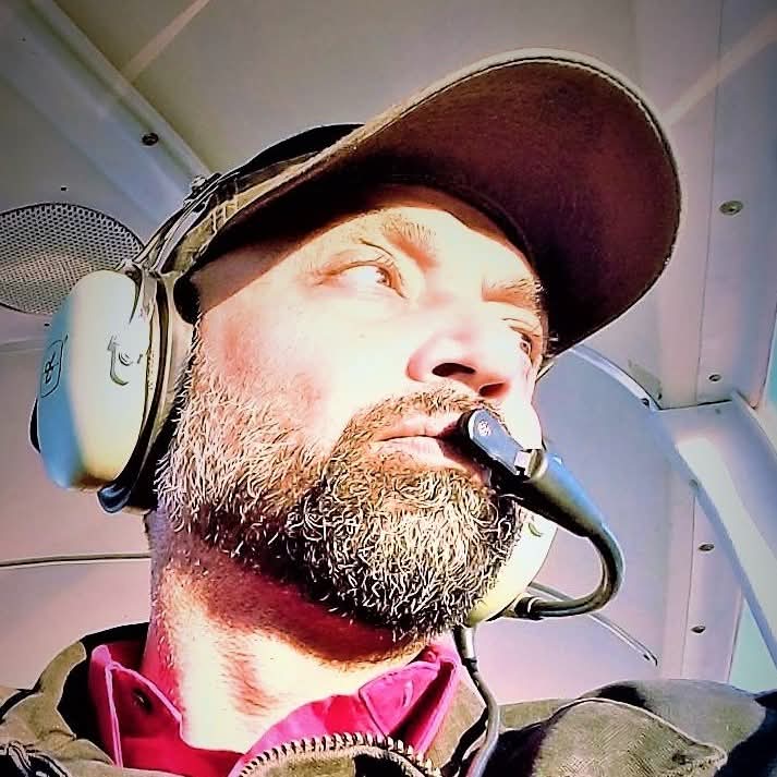 A man with a beard wearing a cap and headset looks out from the cockpit of an aircraft, sunlight illuminating his face—a scene reflecting the mentor protégé flight instruction approach that shapes confident aviators. - CFI Academy