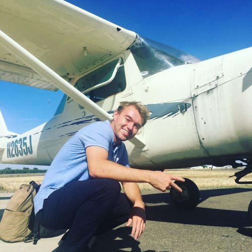 A smiling man in a light blue shirt, likely a Certificated Flight Instructor, crouches beside a small white airplane on a sunny day, with a backpack on the ground nearby and clear blue sky in the background. - CFI Academy