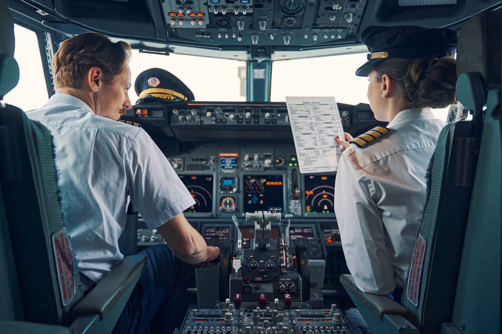 Two airline pilots in uniform, including a Certificated Flight Instructor, sit in the cockpit. One holds a flight checklist as they both study the controls and instruments, preparing for departure. - CFI Academy