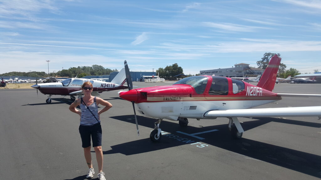A woman in sunglasses, a striped tank top, and shorts stands on an airport tarmac near a small red and white airplane at a flight school California, with another plane and buildings in the background under a clear blue sky. - CFI Academy