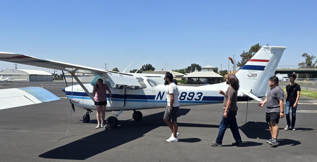 Five people walk around a small white and blue single-engine airplane parked on an airport tarmac under a clear sky, discussing flight training cost. Hangars and trees are visible in the background. - CFI Academy