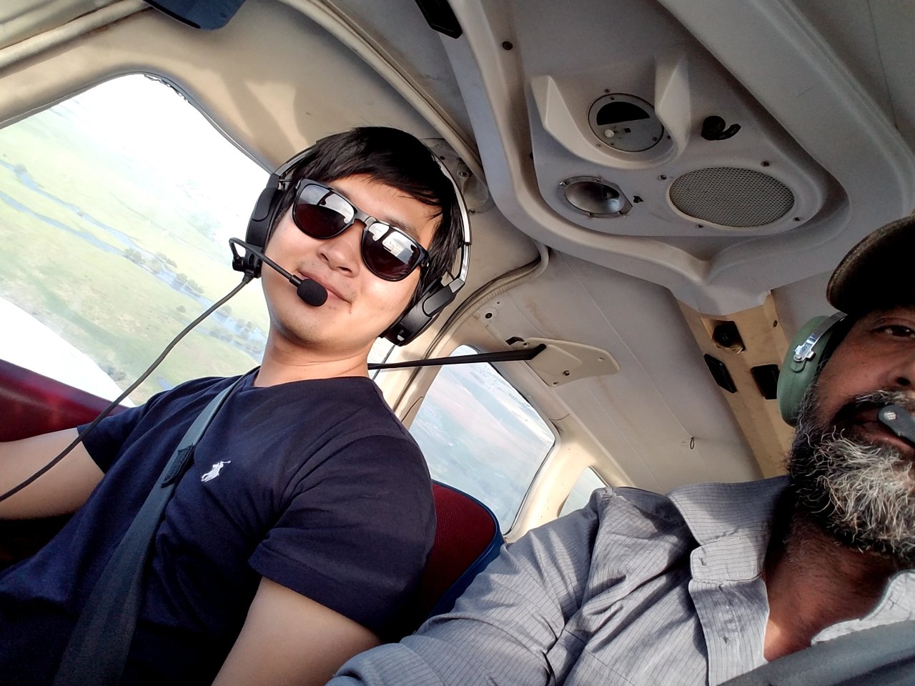 Two men wearing headsets sit inside a small airplane cockpit during a flight instructor program. The man on the left, in sunglasses and a black shirt, is in the pilot seat, smiling. Outside the window, scenic views reward their journey to become a pilot. - CFI Academy