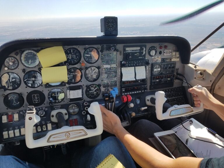 Two people sit in a small airplane cockpit, each holding a control yoke. The dashboard is filled with various flight instruments, gauges, and switches. Notepads and checklists are visible on their laps and on the panel. - CFI Academy